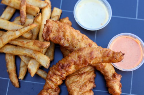 Crispy fried chicken tenders with two dipping sauces, accompanied by golden French fries on a gray surface.