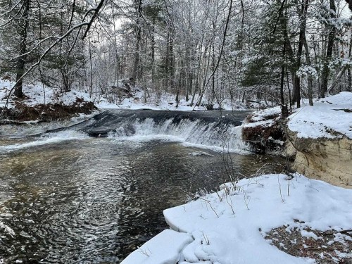A serene winter scene featuring a small waterfall flowing into a calm river, surrounded by snow-covered trees.