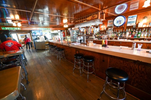 A cozy bar interior with a long counter, stools, and shelves of liquor, featuring wooden decor and a TV in the background.