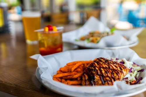A plate with grilled salmon, sweet potato fries, and coleslaw, with drinks in the background.