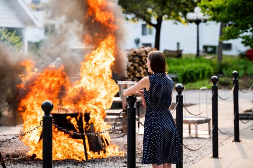 A woman in a polka dot dress stands near a fire, observing flames and smoke in a park setting.