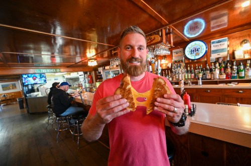 A man with a beard holds a grilled cheese sandwich, showcasing its melted cheese, in a bar setting.