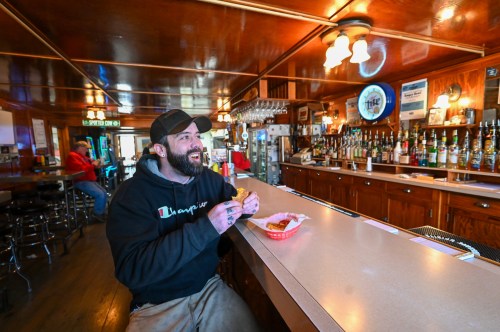 A man in a black hoodie smiles while sitting at a bar, enjoying food in a cozy, wooden-paneled restaurant.