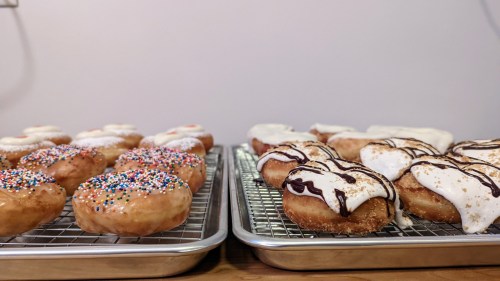 Two trays of donuts: one with colorful sprinkles and the other topped with white icing and chocolate drizzle.
