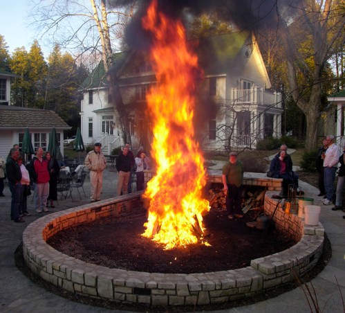 A large bonfire blazes in a stone pit, surrounded by people watching in a backyard setting.