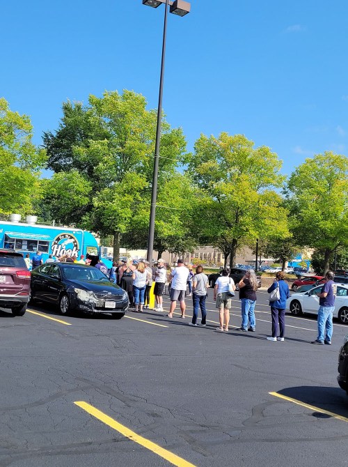 A long line of people waiting near a food truck in a parking lot, surrounded by trees and clear blue skies.