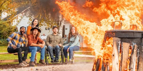 A group of five people sitting by a large bonfire, surrounded by trees and a cozy outdoor setting.