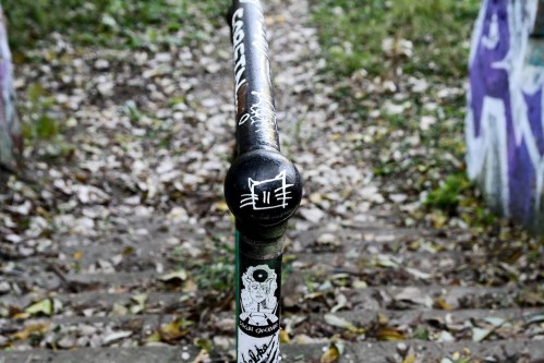 A close-up of a black railing with a cat sticker, surrounded by fallen leaves and graffiti on nearby walls.