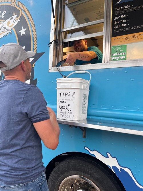 A customer at a food truck interacts with the vendor, who is smiling and handing over food. A tip bucket is visible.
