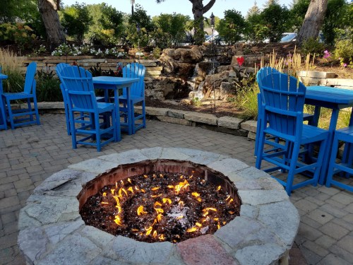 A cozy outdoor seating area with blue chairs around a stone fire pit, featuring flames and a small waterfall in the background.
