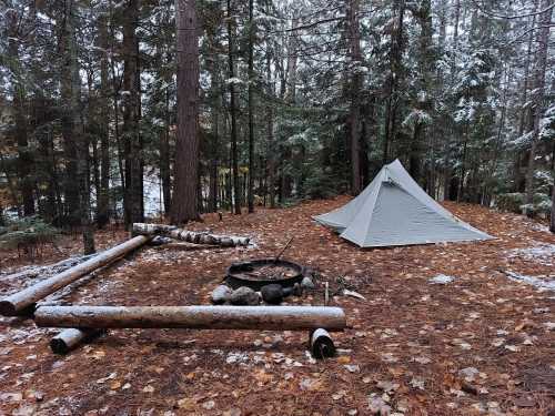 A tent set up in a snowy forest clearing, with a fire pit and logs nearby, surrounded by tall trees and fallen leaves.