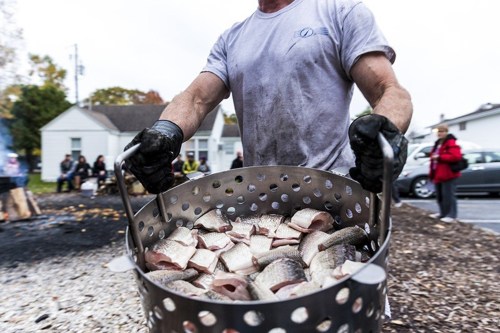 A person carries a basket of fish, with a crowd in the background, during an outdoor cooking event.