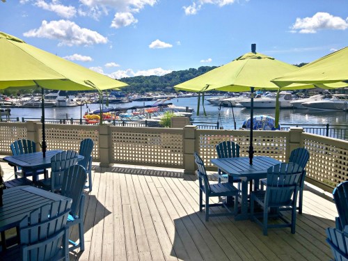 A sunny marina view with green umbrellas over blue tables, boats docked in the background, and a clear blue sky.