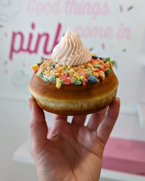 A hand holding a donut topped with pink frosting and colorful cereal bits, against a pink wall with text.