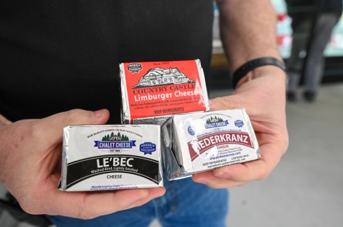 A person holding three types of cheese packages: Limburger, Le'Bec, and Nederkranz, all from Chalet Cheese.