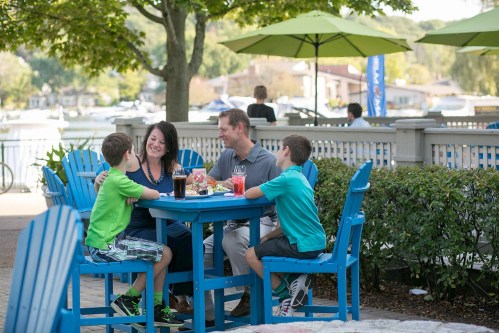 A family enjoys a meal at an outdoor table, surrounded by greenery and water, with drinks and food on the table.