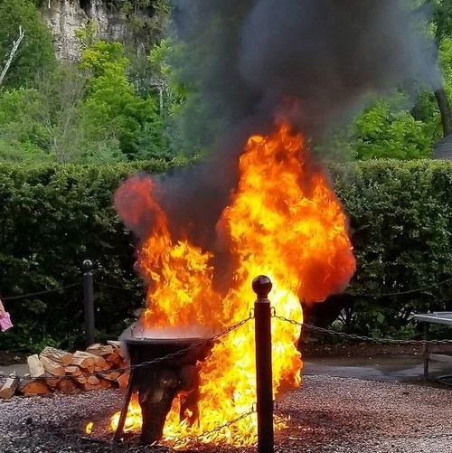 A large fire with thick black smoke billowing from a metal container, surrounded by greenery and firewood.