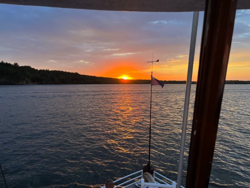 Sunset over calm waters, viewed from a boat with a flag and trees lining the horizon.