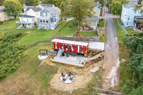 Aerial view of a red train car converted into a home, surrounded by greenery and nearby houses. Outdoor seating area visible.