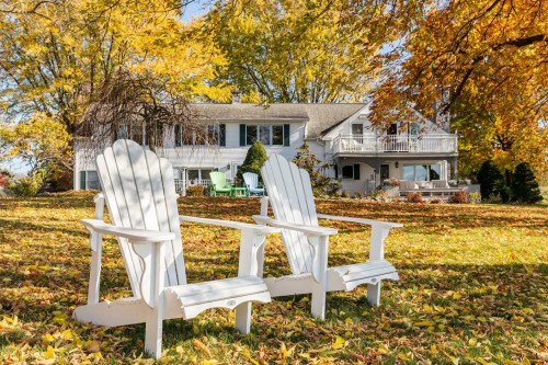 Two white Adirondack chairs sit on a lawn covered in autumn leaves, with a house and colorful trees in the background.