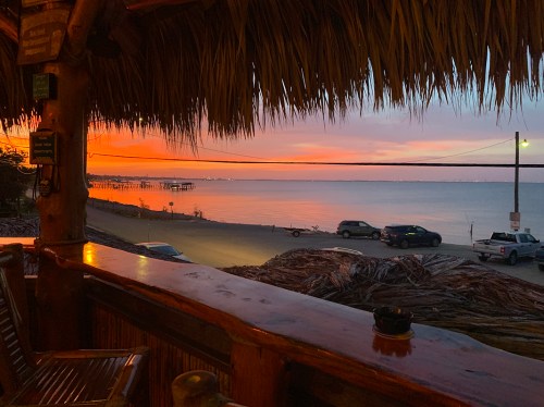 A sunset view over a calm sea from a rustic wooden deck, with silhouettes of cars and a pier in the distance.