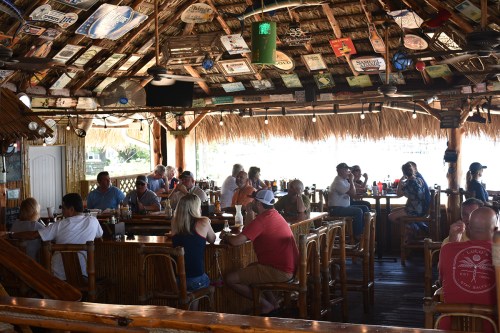 A lively bar with a thatched roof, filled with patrons enjoying drinks and conversation at wooden tables.