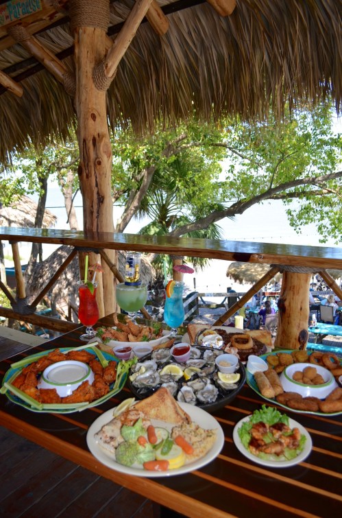 A vibrant outdoor table set with a variety of seafood dishes, appetizers, and colorful cocktails, with a beach view.