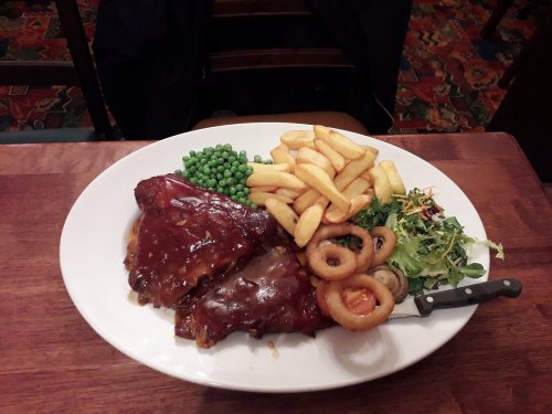 A plate of BBQ ribs with sauce, fries, green peas, salad, and onion rings on a wooden table.