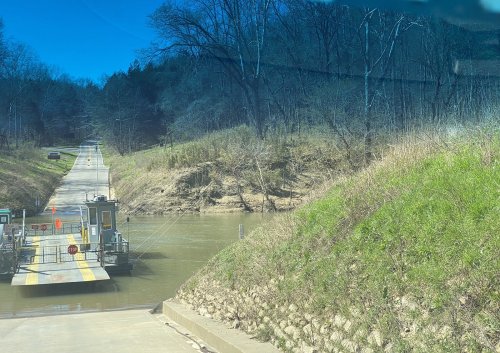 A ferry docked at a riverbank, with a road leading to a wooded area under a clear blue sky.