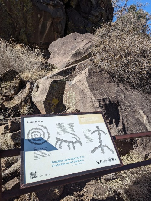 A sign detailing petroglyphs on a rocky surface, surrounded by sparse vegetation and a clear blue sky.