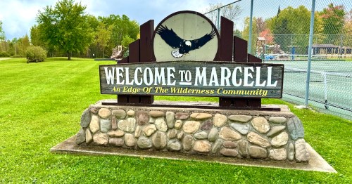 A brown wooden sign on a fieldstone pedestal depicts a bald eagle and reads "Welcome to Marcell."