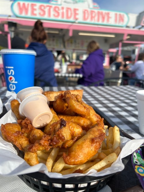 A basket of fried food with dipping sauce and fries, set on a checkered tablecloth at Westside Drive-In.