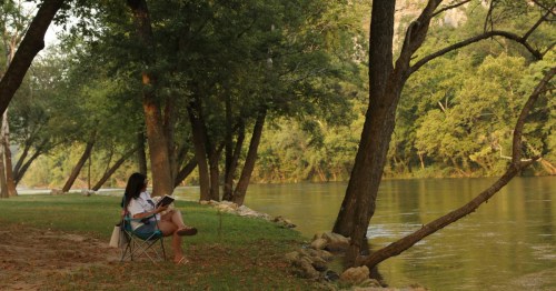 a woman reading a book sits in a chair by the river at white buffalo resort in arkansad