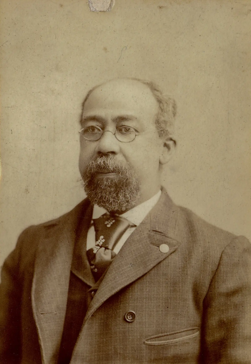 A sepia-toned portrait of a man with glasses, a beard, and a suit, looking directly at the camera.