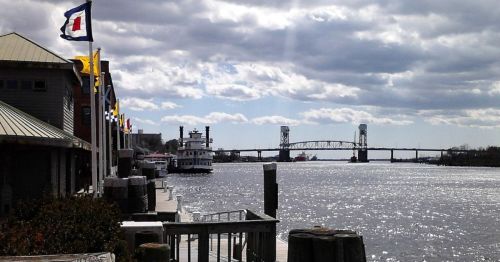 A scenic view of a river with boats, flags, and a bridge under a partly cloudy sky.