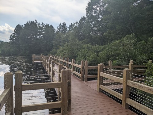 A wooden boardwalk extends over calm water, surrounded by lush greenery and trees under a cloudy sky.