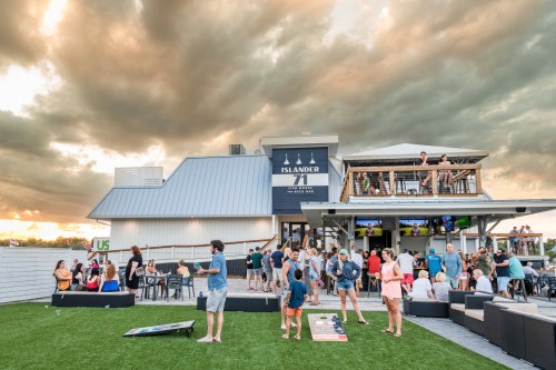 A lively outdoor scene at Islander 71, with people socializing, playing games, and enjoying a cloudy sunset.