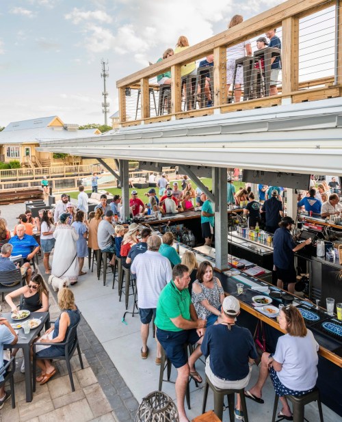 A lively outdoor dining scene with people enjoying food and drinks at a restaurant with a rooftop seating area.