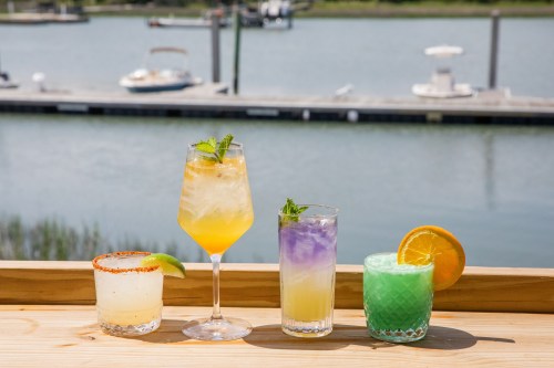Four colorful cocktails on a wooden railing, with a scenic waterfront view in the background.