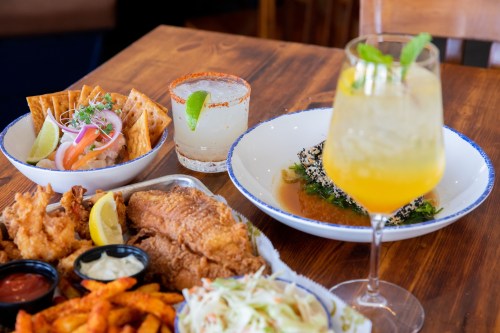 A vibrant spread of fried fish, tacos, coleslaw, and colorful cocktails on a wooden table.