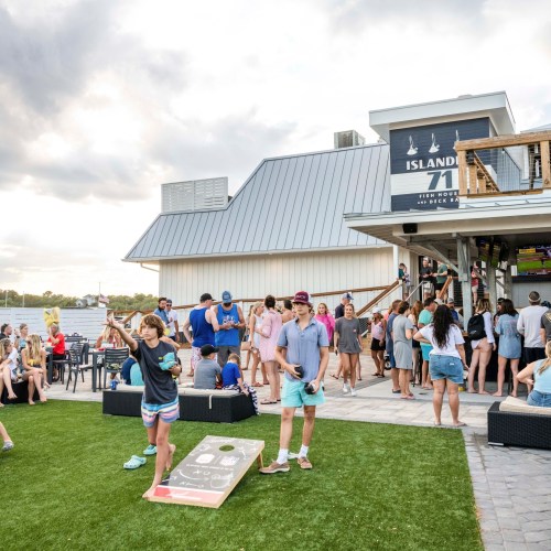 A lively outdoor scene at a waterfront venue with people playing cornhole and socializing on a grassy area.