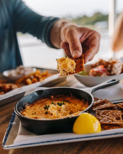 A hand dips a cracker into a creamy dip served in a small skillet, with lemon and snacks on the table.