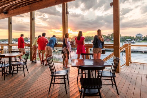 People enjoying a sunset view from a wooden deck, with tables and chairs in the foreground.