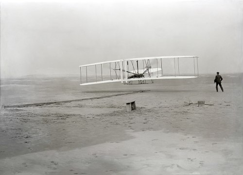 A historic black-and-white photo of the Wright brothers' airplane in flight, with a man observing from the ground.