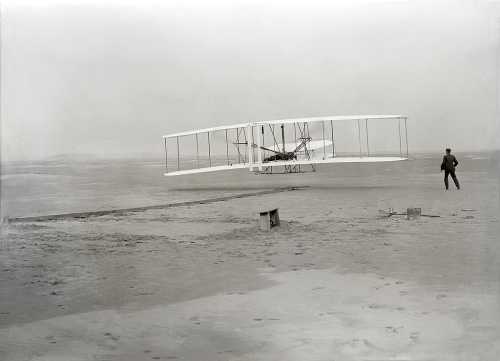 A historic black-and-white photo of the Wright brothers' airplane in flight, with a man observing from the ground.