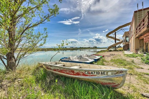 A weathered boat rests on the shore by a calm lake, with a waterslide and buildings in the background under a blue sky.