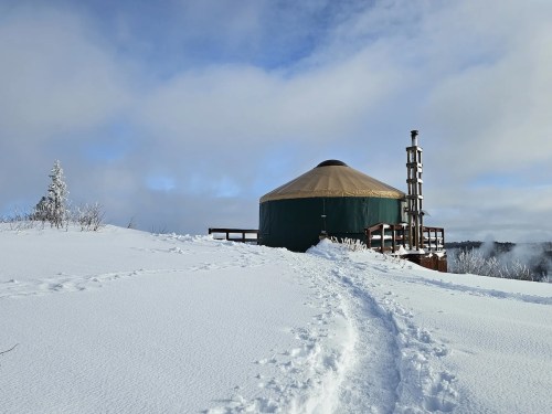 A yurt on a snowy landscape, surrounded by trees and under a cloudy sky, with a path leading to it.