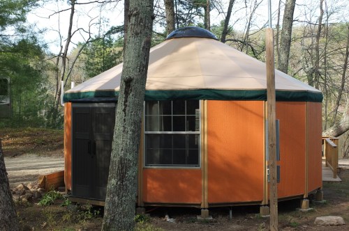 A round, orange yurt with a green roof, set among trees in a wooded area.