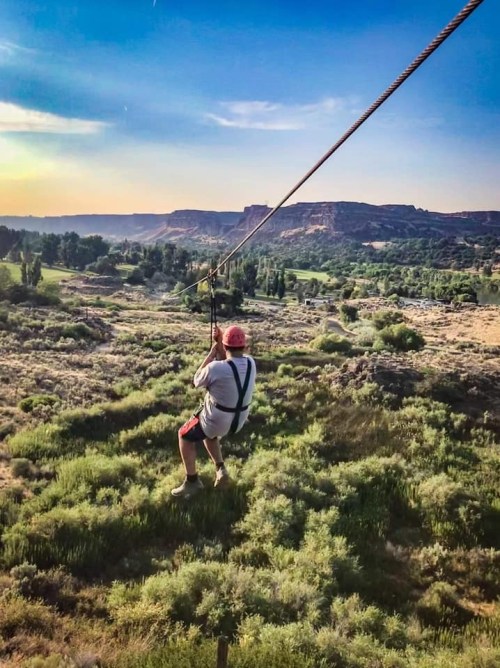 A person zip-lining over a lush green landscape with hills in the background during sunset.