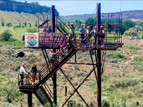 A group of people in helmets stands on a zip line platform, smiling and waving, surrounded by greenery and hills.